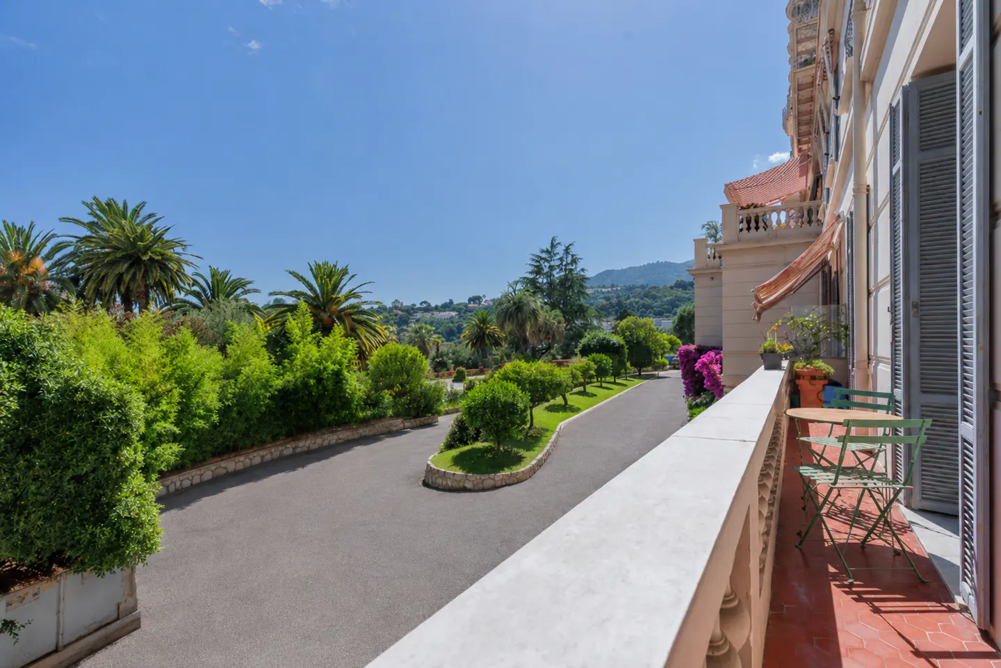 View from a balcony overlooking a paved driveway, lush green palm trees, and distant hills under a clear blue sky.
