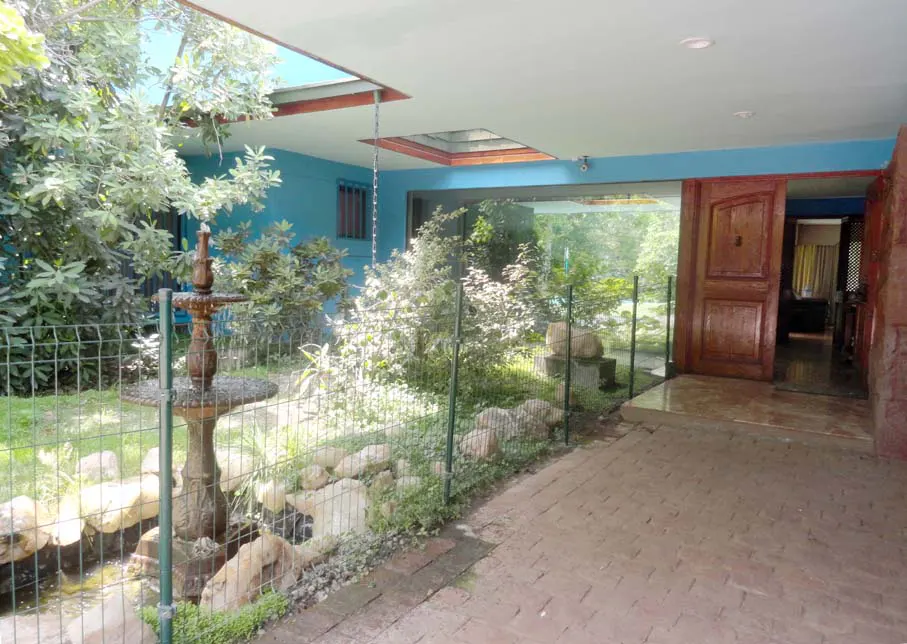 Exterior view of a blue house with a water fountain, greenery, and a brick walkway leading to an open wooden door.