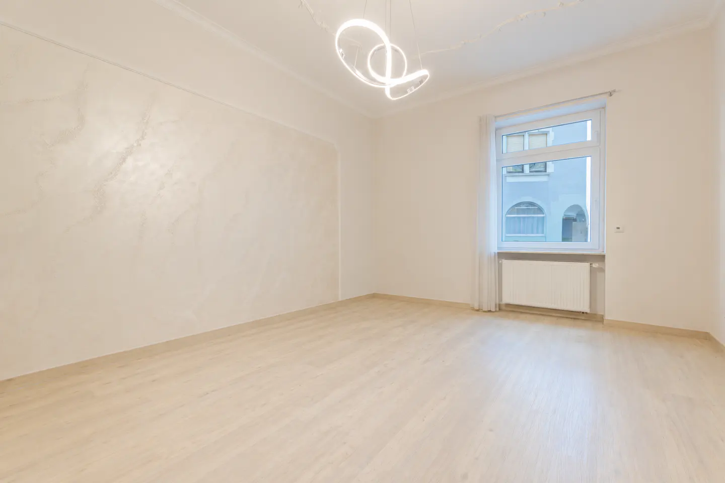 Bright, empty room with light wood floors, white walls, and a modern, looping light fixture. A window with sheer curtains overlooks a building.