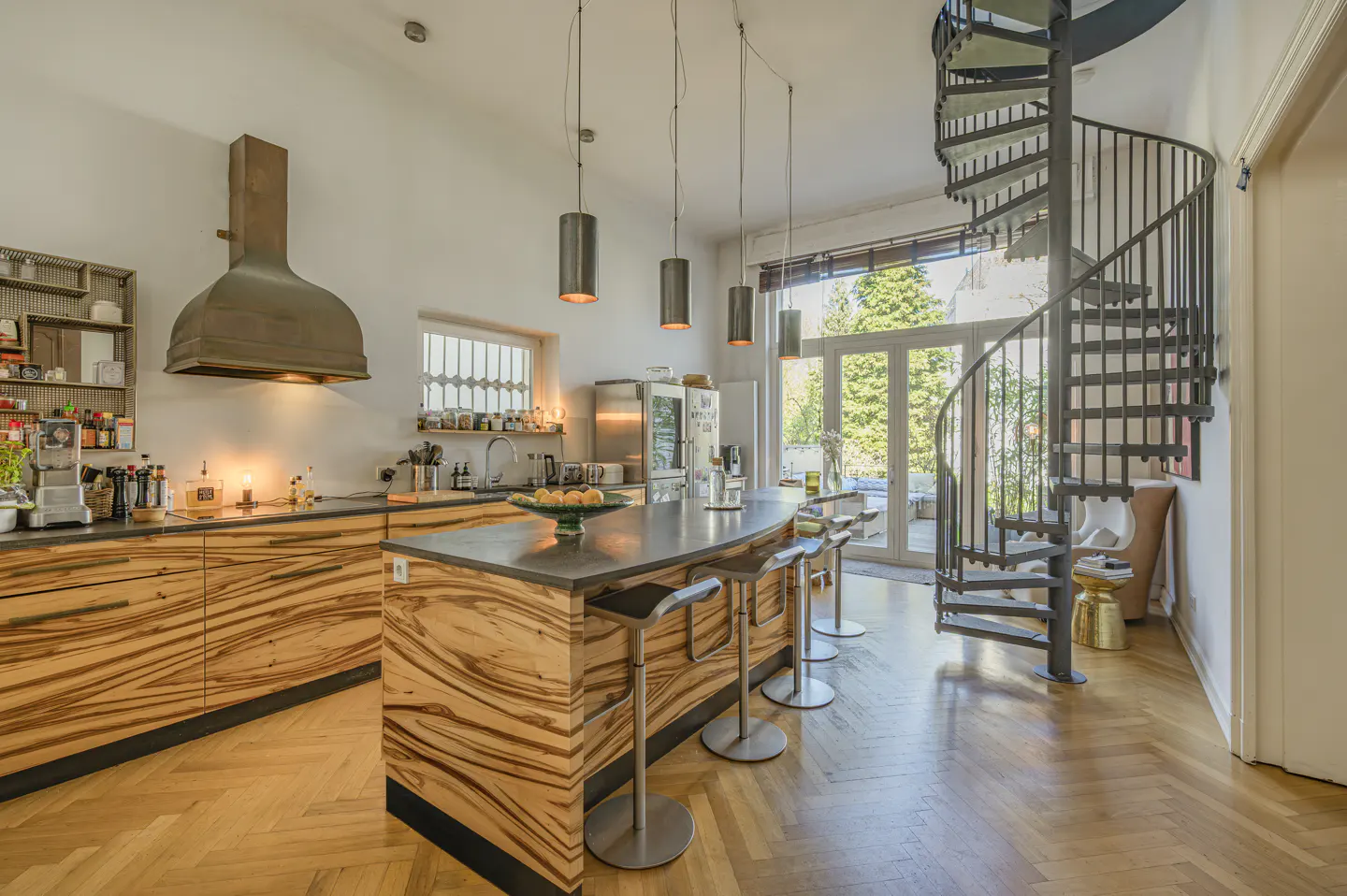 A modern kitchen with wood cabinets, a gray island with stools, and a spiral staircase.