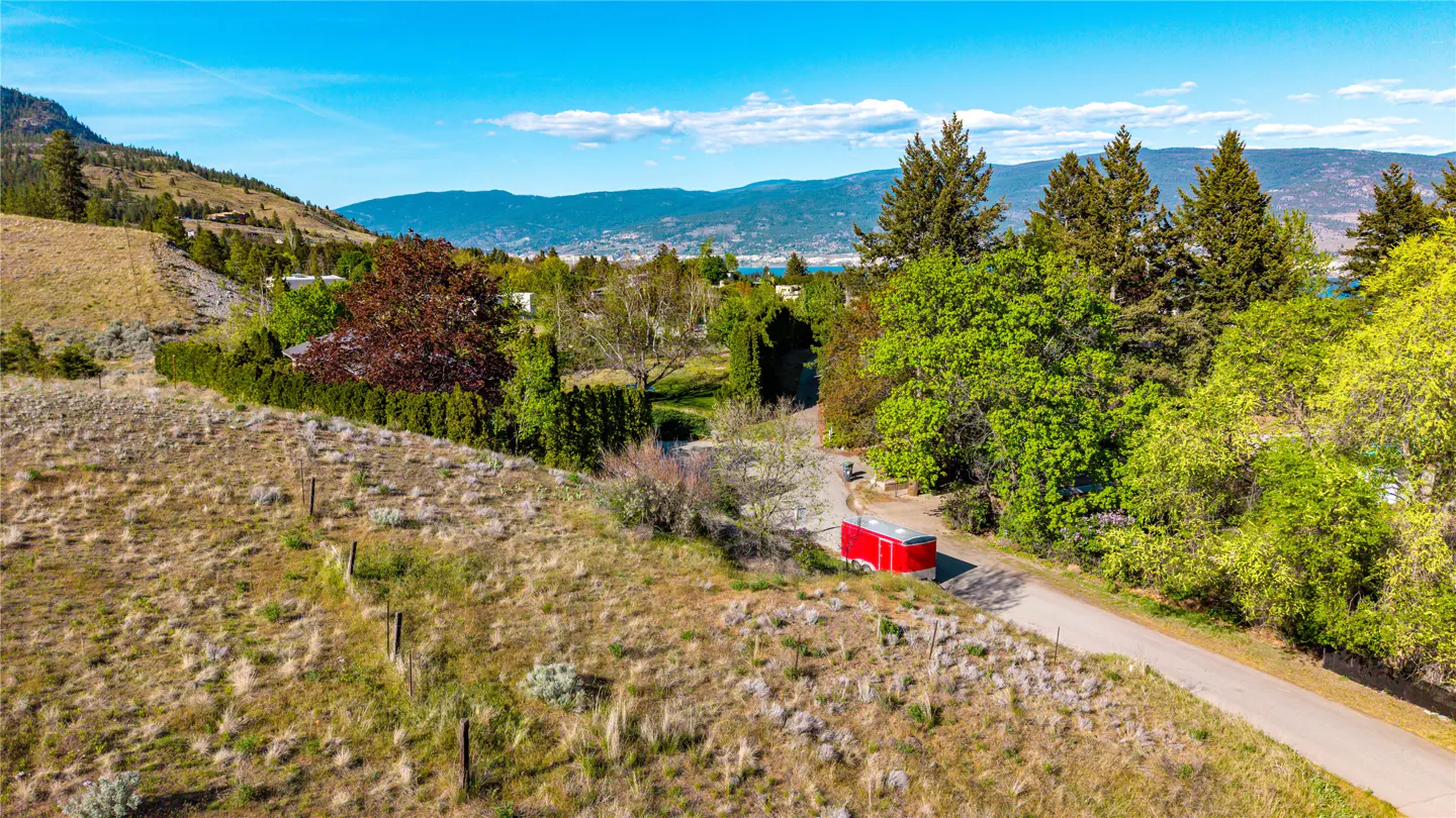 Aerial view of a property with a red trailer on a road, surrounded by trees and a grassy hillside. Mountains and a blue sky are in the background.