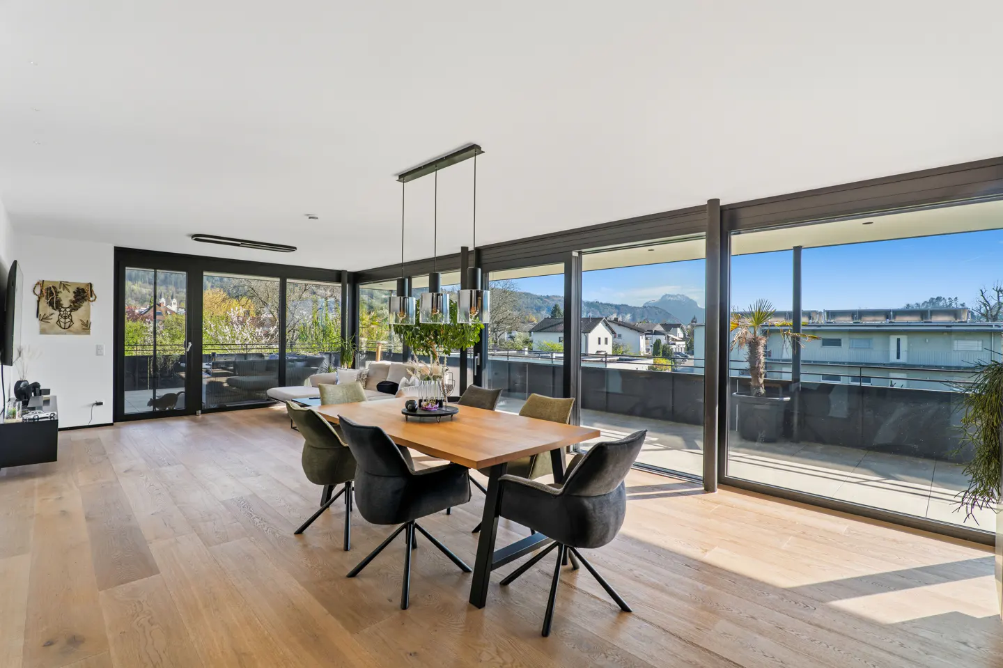 Bright, open-plan living and dining area with wood floors, large windows, and a view of the mountains. A wooden table with chairs is in the foreground.
