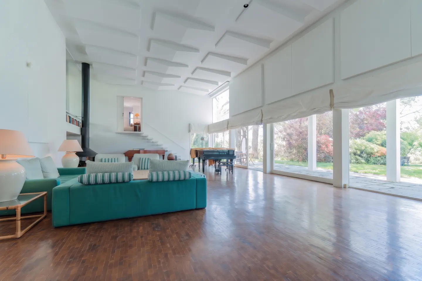 Bright, modern living room with a teal sofa, piano, fireplace, and floor-to-ceiling windows overlooking a green yard.