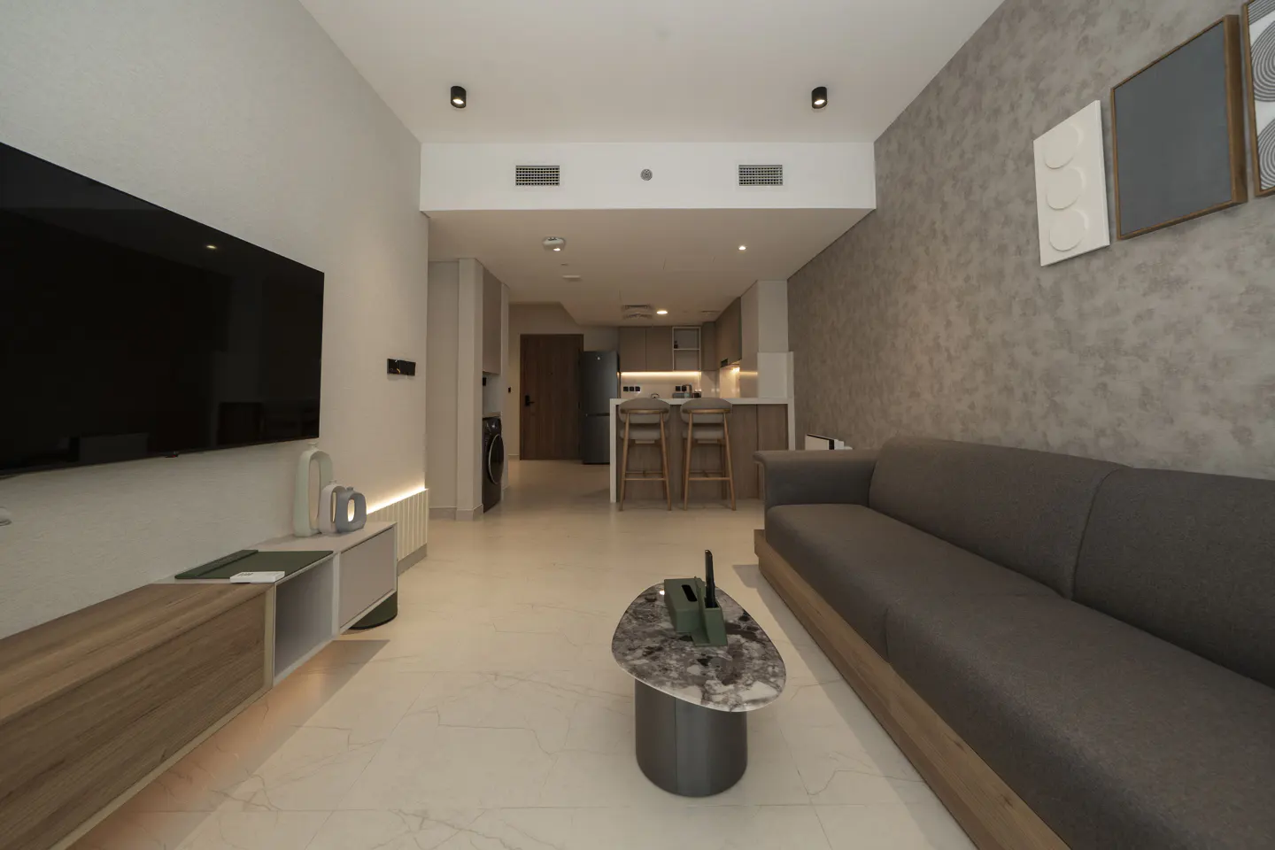 A modern apartment interior with a gray sofa, marble coffee table, TV, and open kitchen.