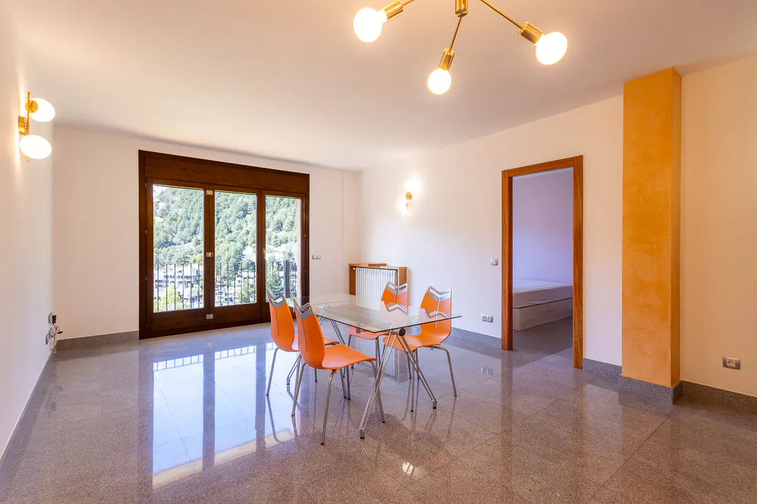 Bright, modern dining room with orange chairs, glass table, and a view of green hills through a large window.