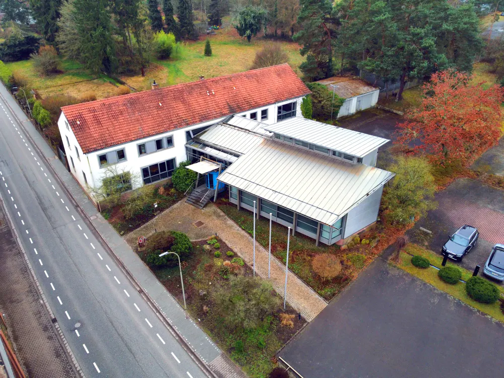 Aerial view of a two-story white building with a red tile roof, connected to a modern, single-story structure with a silver roof. Cars are parked nearby.
