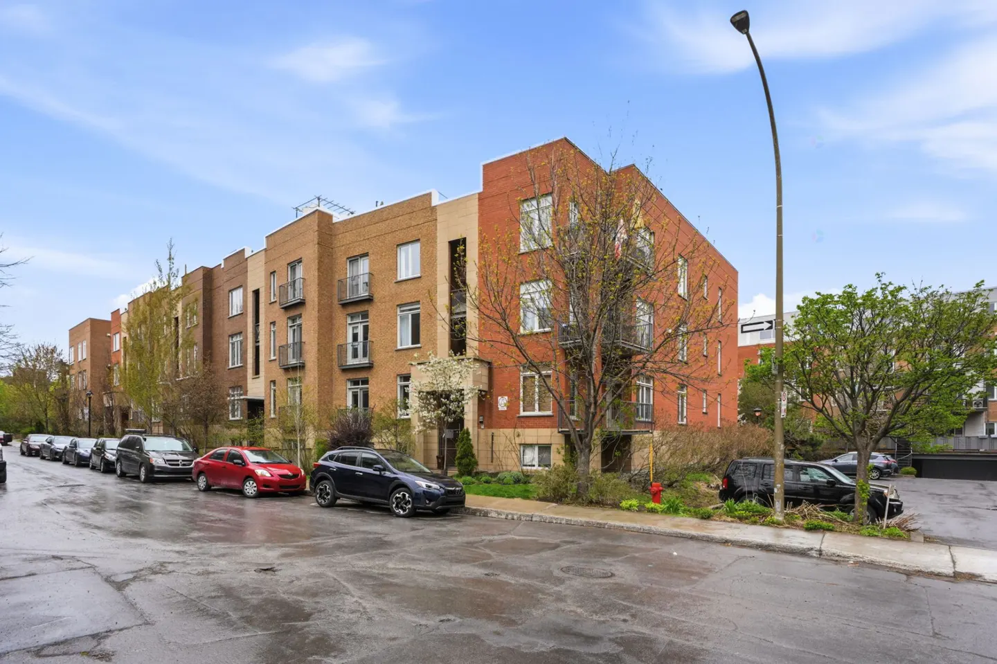 Exterior view of a multi-story brick apartment building with balconies and parked cars on a wet street.