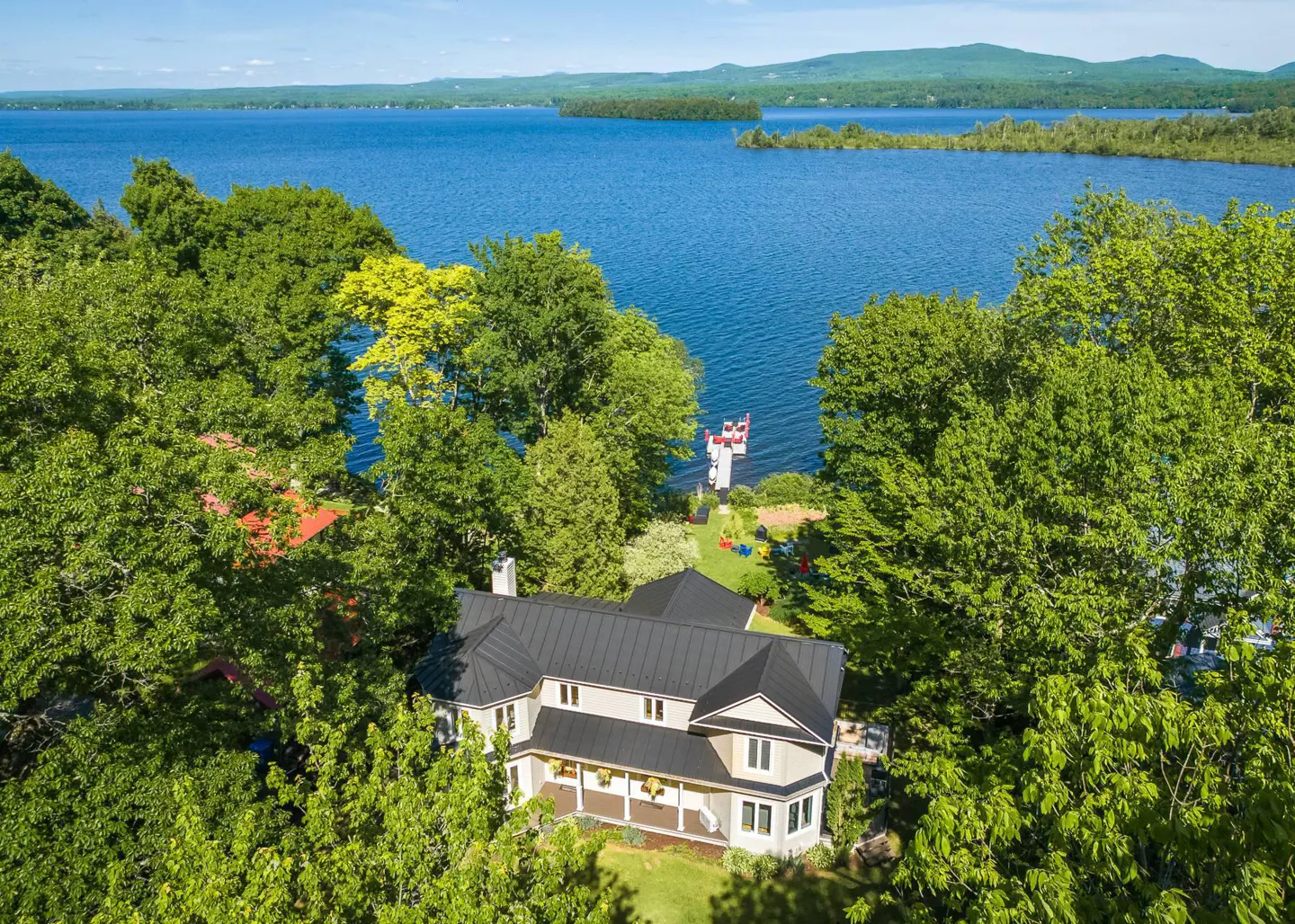 Aerial view of a beige house with a black roof, surrounded by green trees, overlooking a blue lake and mountains.