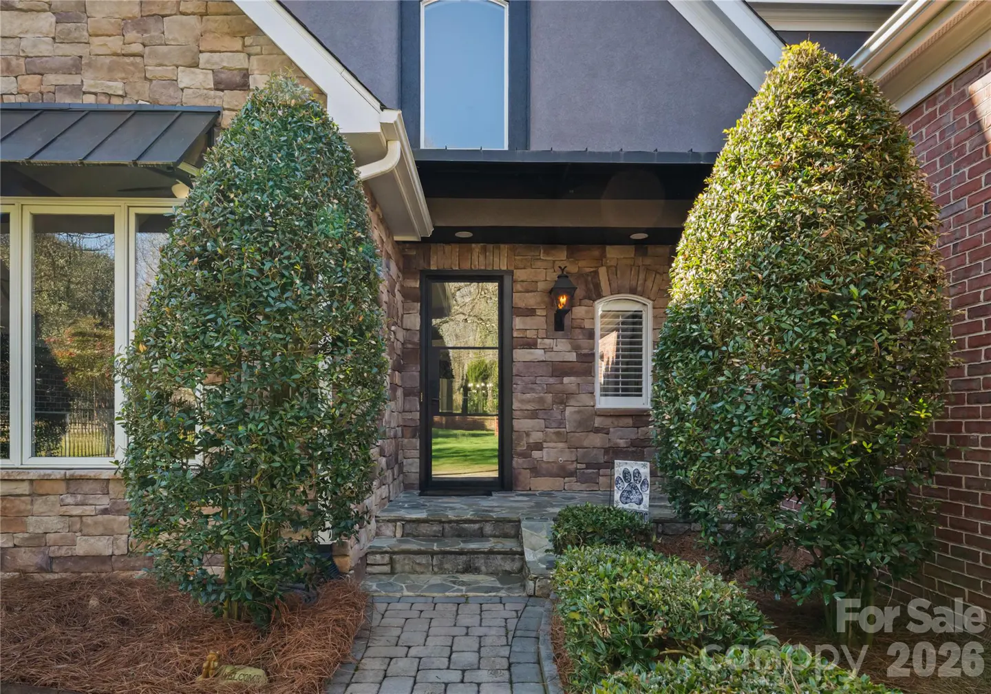 Stone house exterior with a black-framed glass door, flanked by green bushes. A brick walkway leads to the entrance.