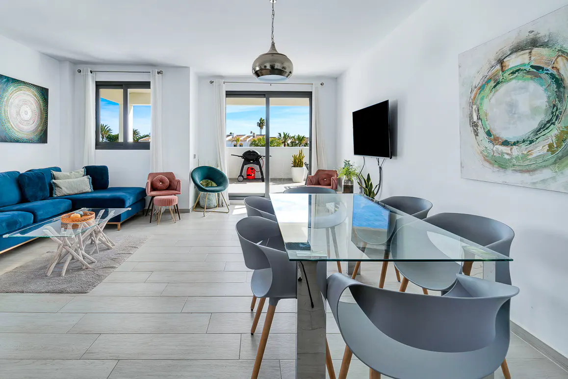 Bright living and dining area with a blue velvet sofa, glass table, and gray chairs. Balcony with grill visible through sliding glass doors.