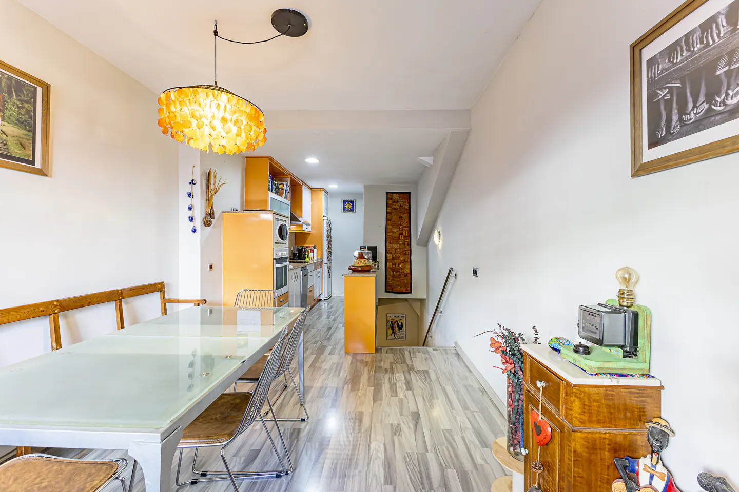 Bright, modern dining room with a long glass table, orange kitchen cabinets, and unique decor. Gray wood floors and white walls.