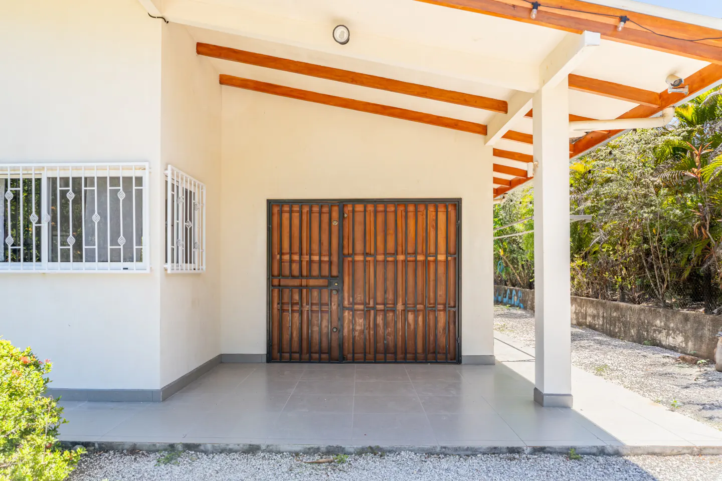 Exterior view of a house with a brown wooden gate, white walls, and barred windows. A covered porch has wooden beams and a gray tiled floor.