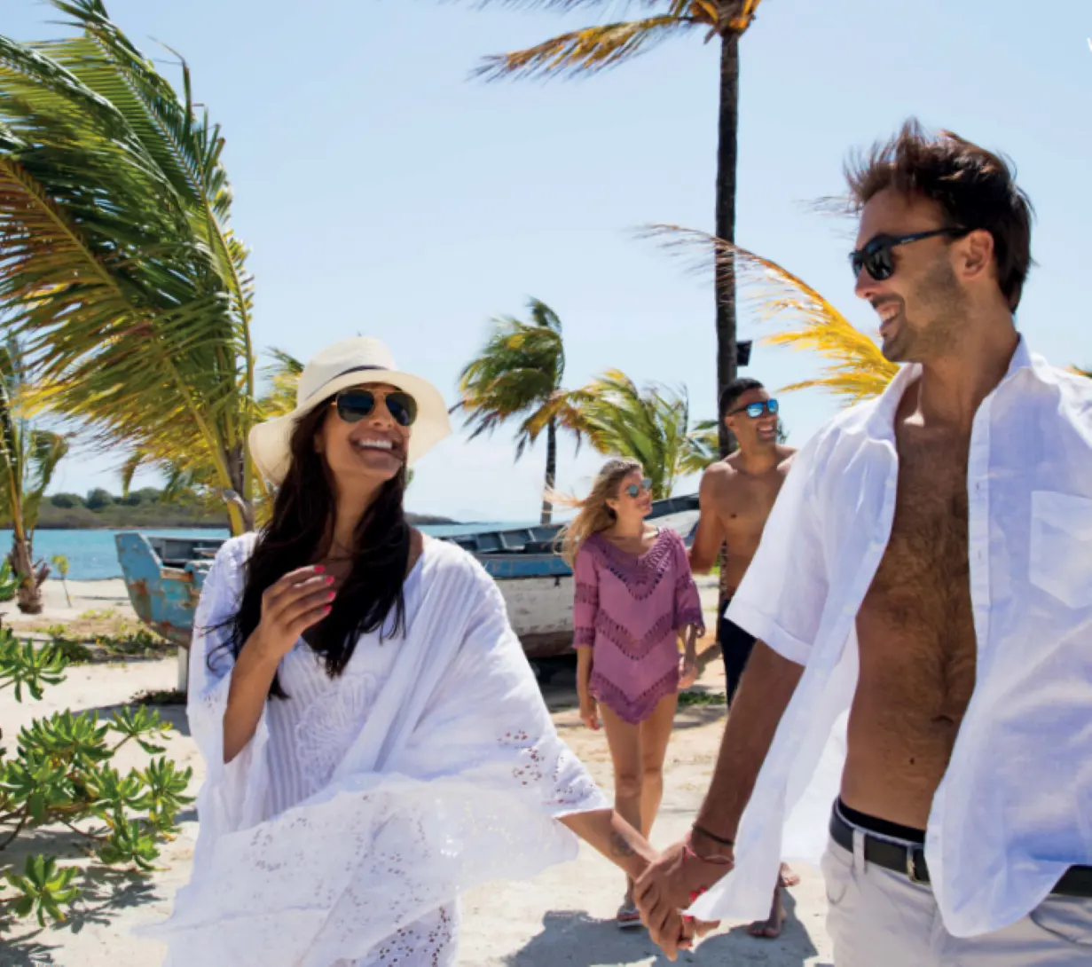 A couple holds hands, walking on a beach with palm trees and blue water. Two other people follow behind them.