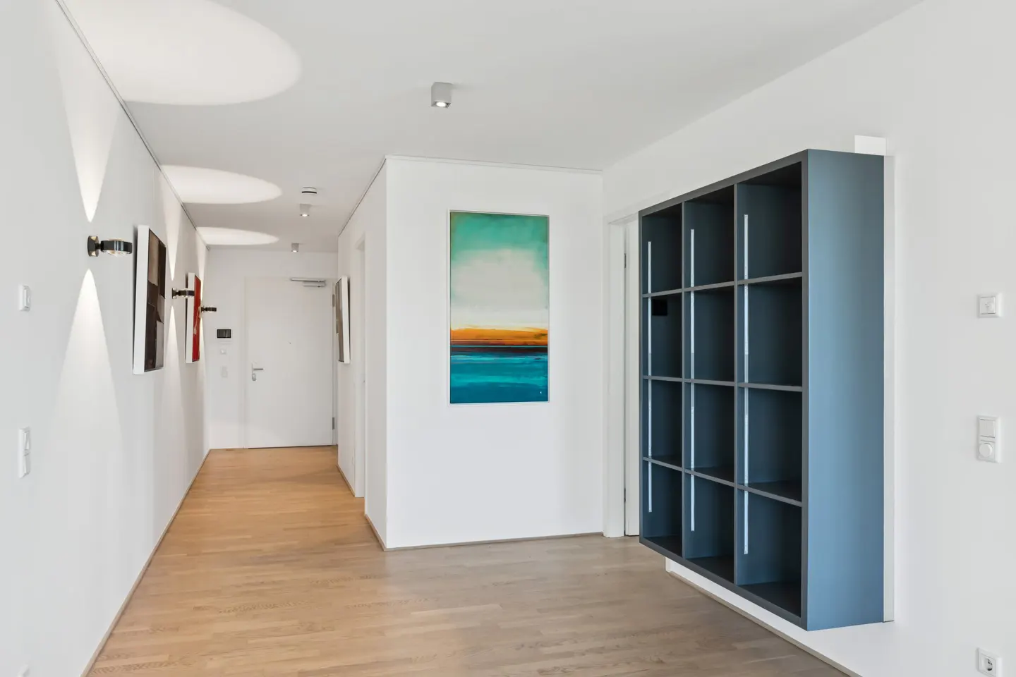 Hallway with light wood floors, white walls, and artwork. A blue shelving unit is mounted on the right wall.