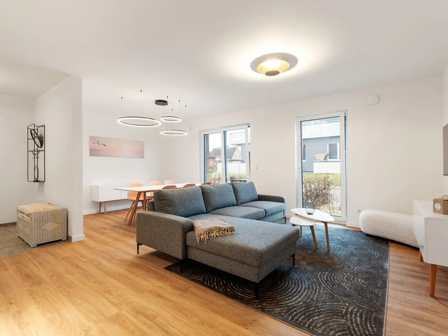 Bright living room with a gray sectional sofa, wood floors, and a patterned rug. A dining table and chairs are visible in the background.