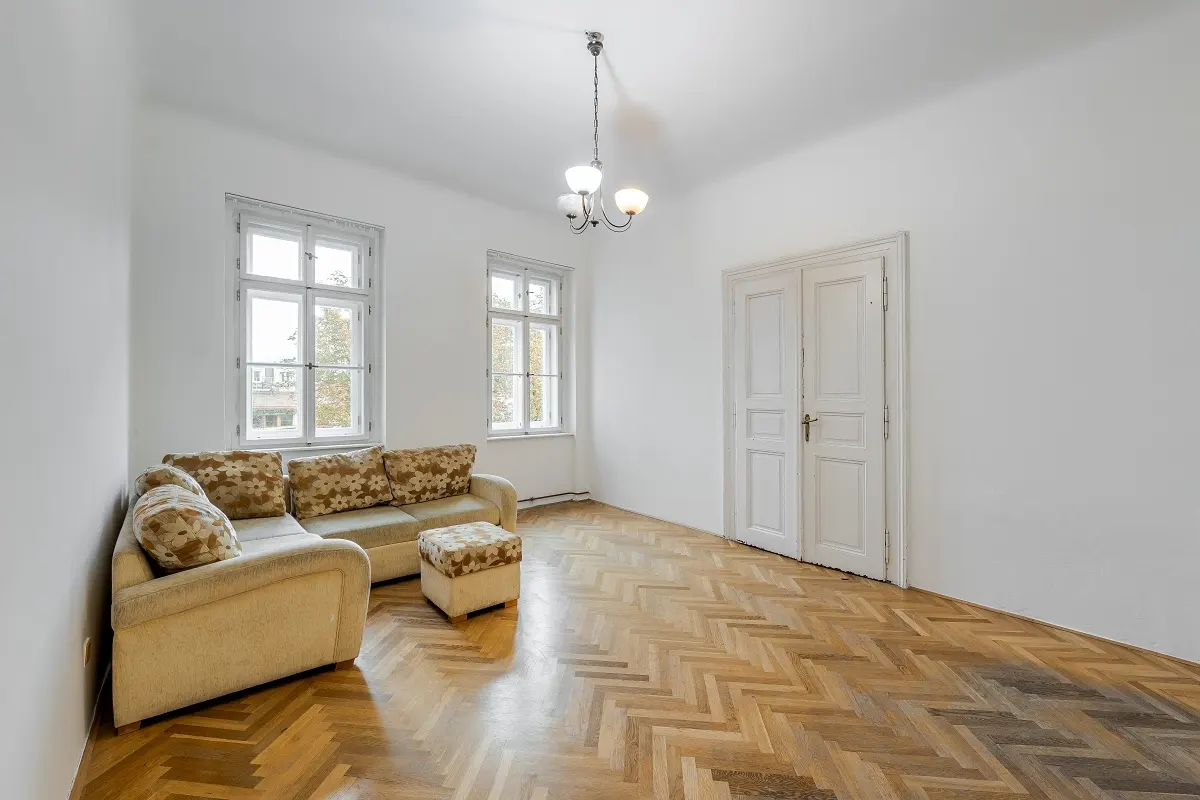 Bright, empty room with herringbone wood floors, white walls, two windows, and a beige sofa with patterned pillows. A white double door is on the right.