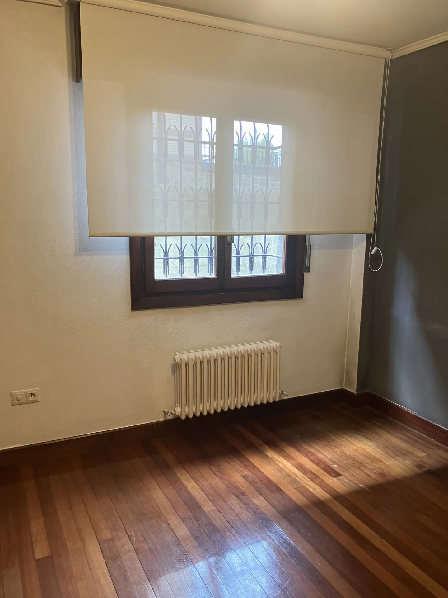 A room with hardwood floors, a window with a white shade, and a radiator.