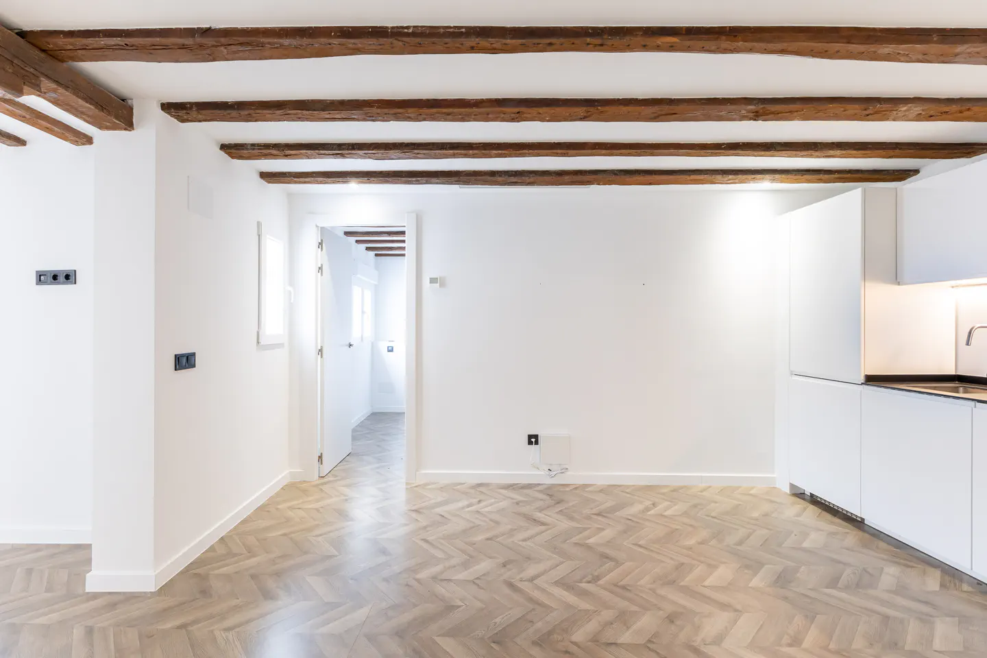 Bright, empty room with white walls, herringbone wood floor, and exposed wood beams. A doorway leads to another room. Modern white kitchen cabinets are visible.