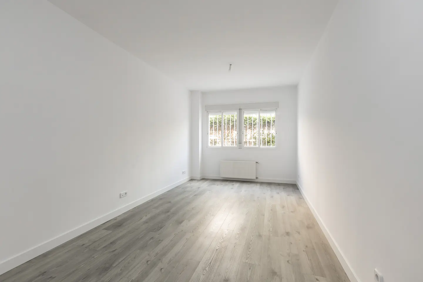 Bright, empty room with white walls and light gray wood floors. A window with white bars looks out onto greenery. A white radiator sits beneath the window.