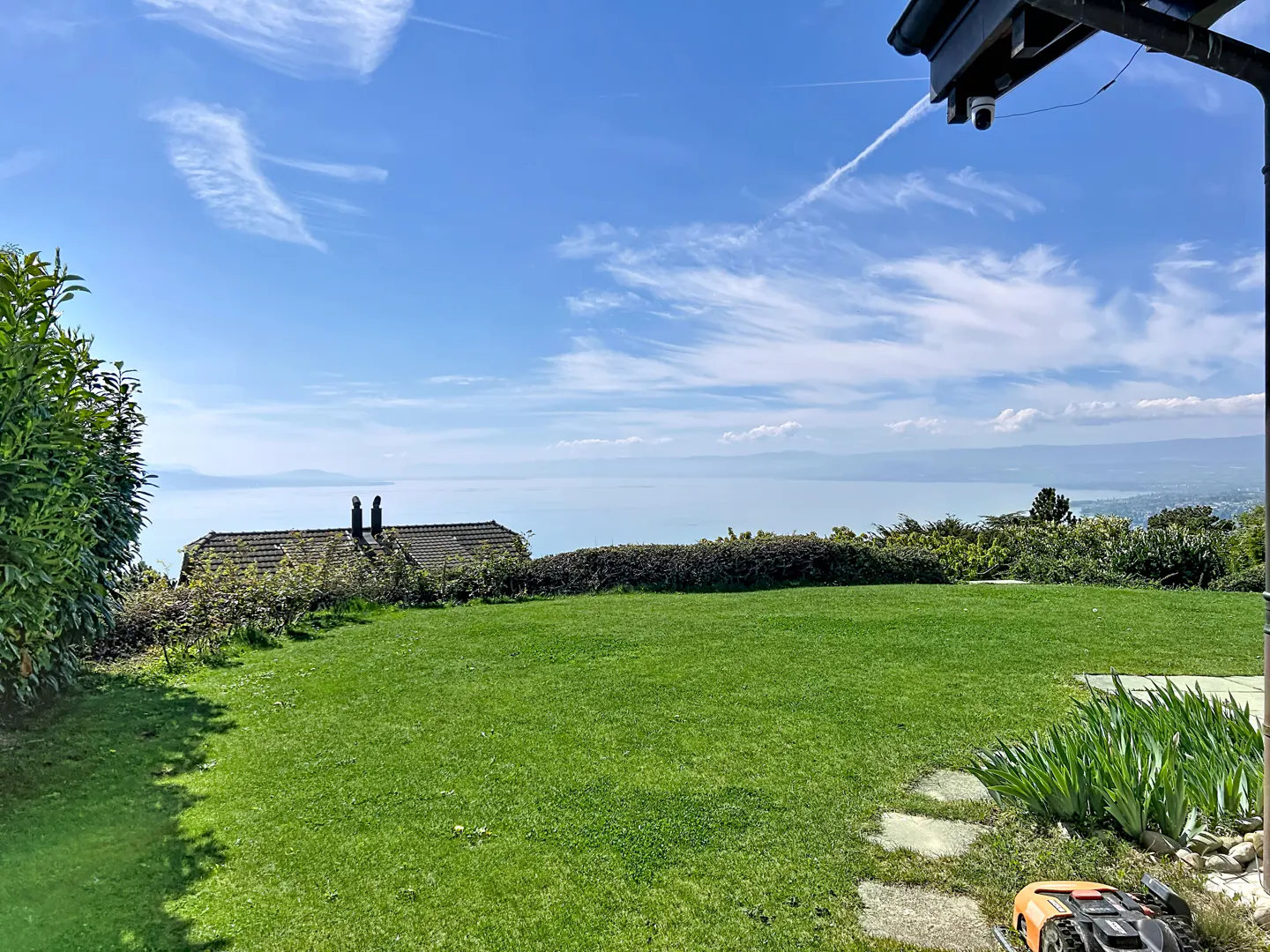 A lush green lawn overlooks a scenic view of a lake and mountains under a blue sky with wispy clouds.