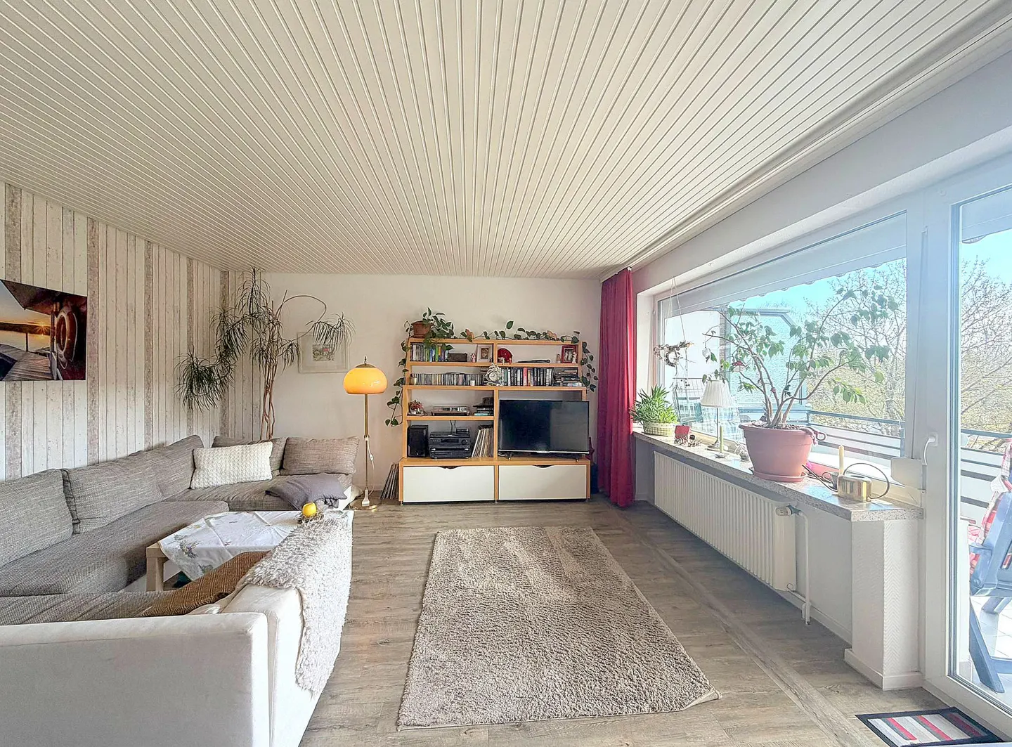 Bright living room with a gray sectional sofa, a large window, and a wooden entertainment center. A beige rug covers the floor.