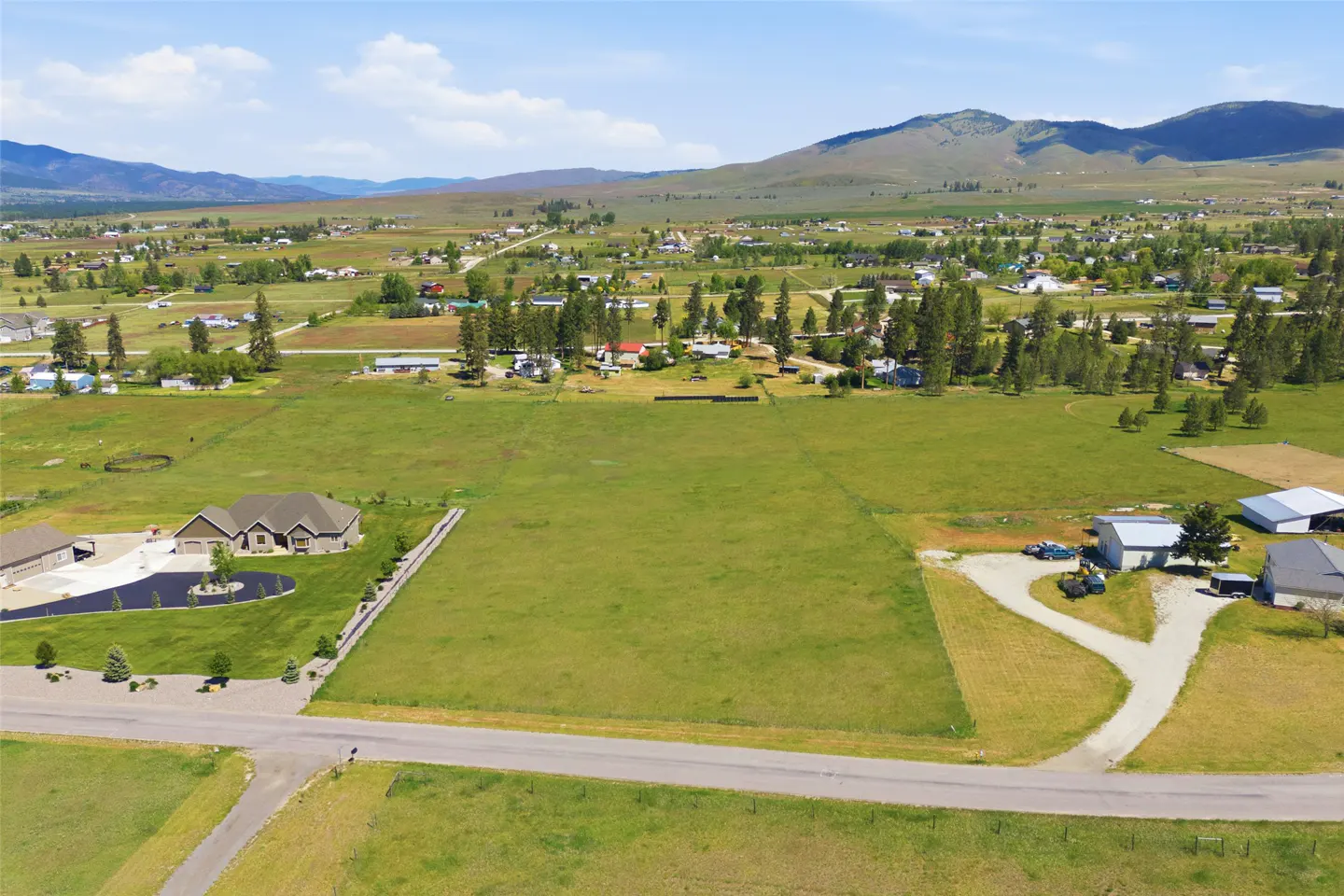 Aerial view of a vacant grassy lot in a rural area with mountains in the background. Houses and trees are scattered throughout the landscape.