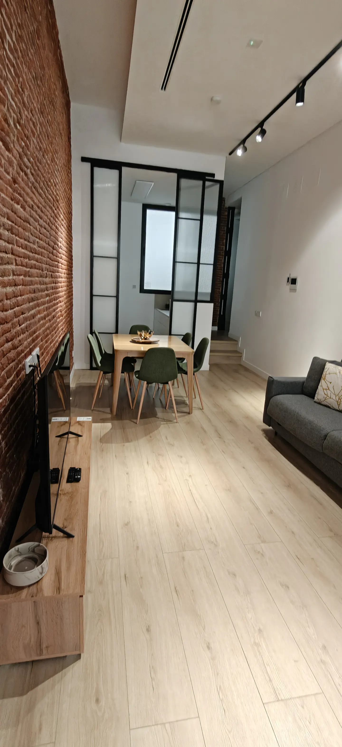 A modern living room with a brick wall, light wood floors, a dining table with green chairs, and a gray sofa.