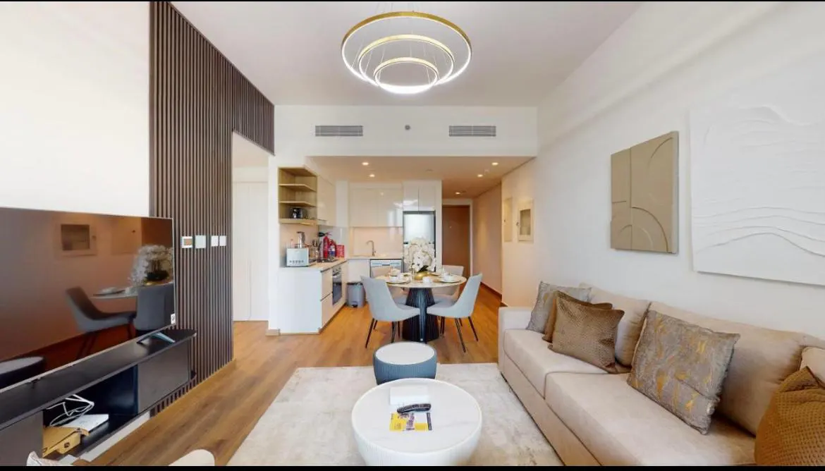 A bright, modern apartment interior with a beige sofa, round table, and kitchen in the background. A large TV is on the left.
