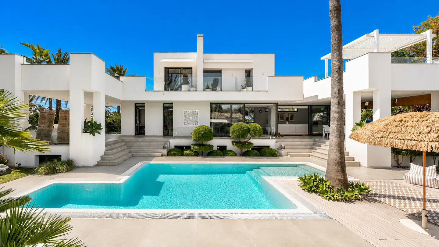 Modern white house with a turquoise pool, palm trees, and a blue sky. The house has large windows and a flat roof.