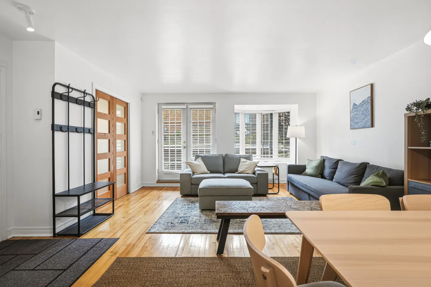Bright living room with hardwood floors, gray sofas, and a dining table. A coat rack stands near a wooden door. Natural light streams through the windows.