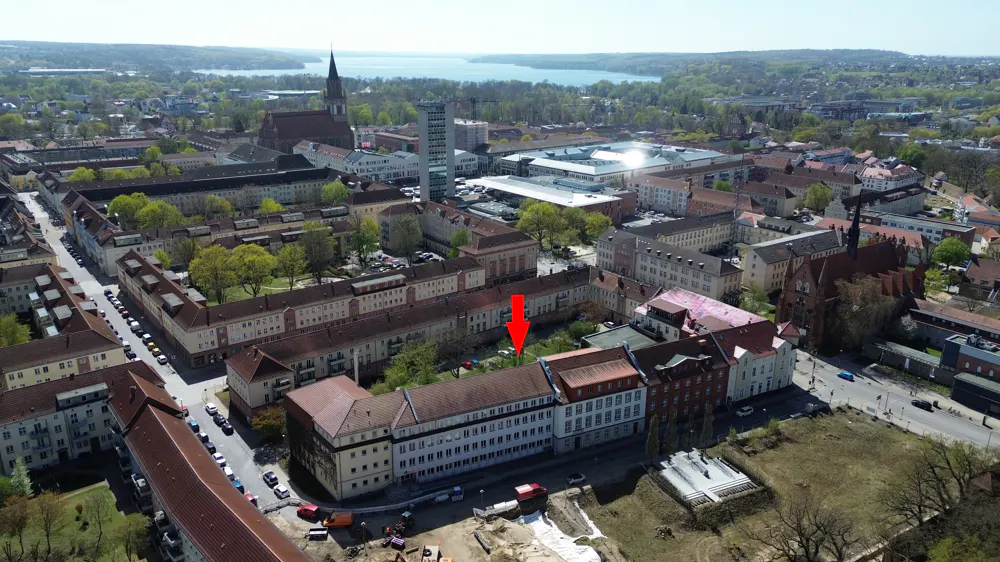 Aerial view of a European city with red arrow pointing to a building with a red roof. A lake is visible in the background.