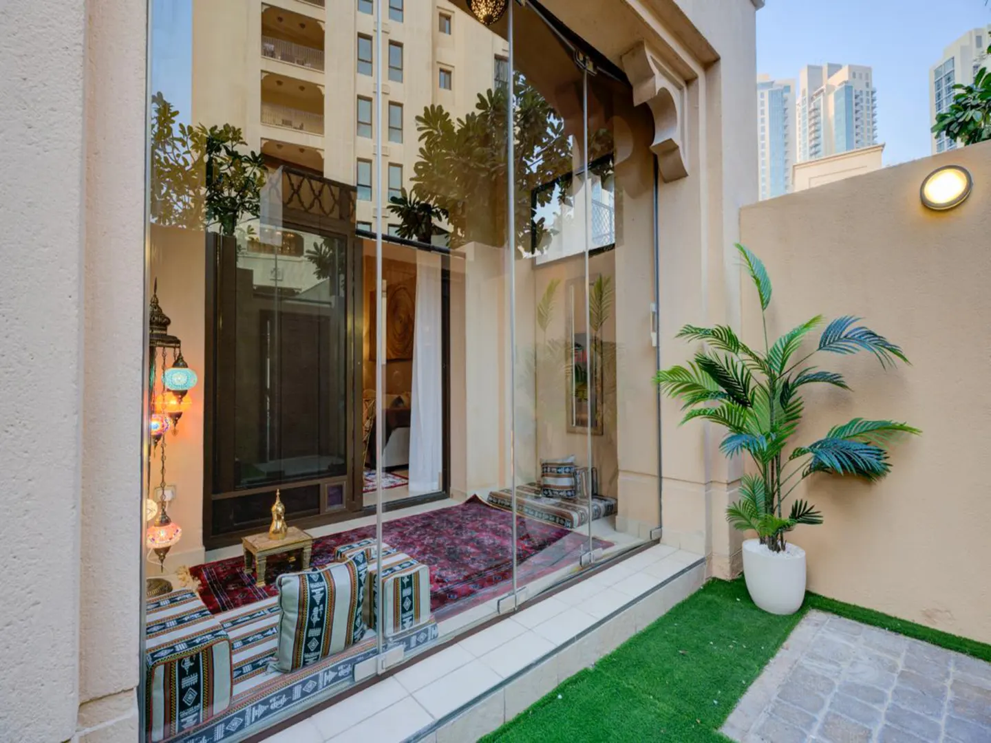 Glass-enclosed patio with a red patterned rug, floor cushions, a decorative lamp, and a potted plant on artificial grass.