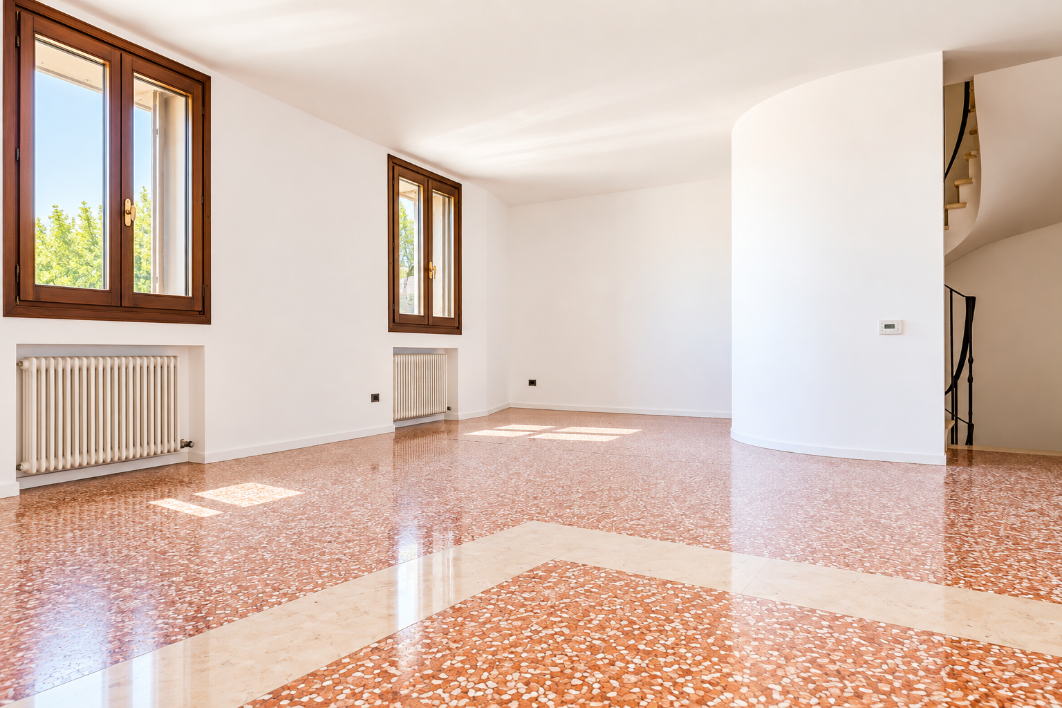 Bright, empty room with white walls, red terrazzo flooring, two wooden windows, radiators, and a curved wall with stairs. [AI-generated text]