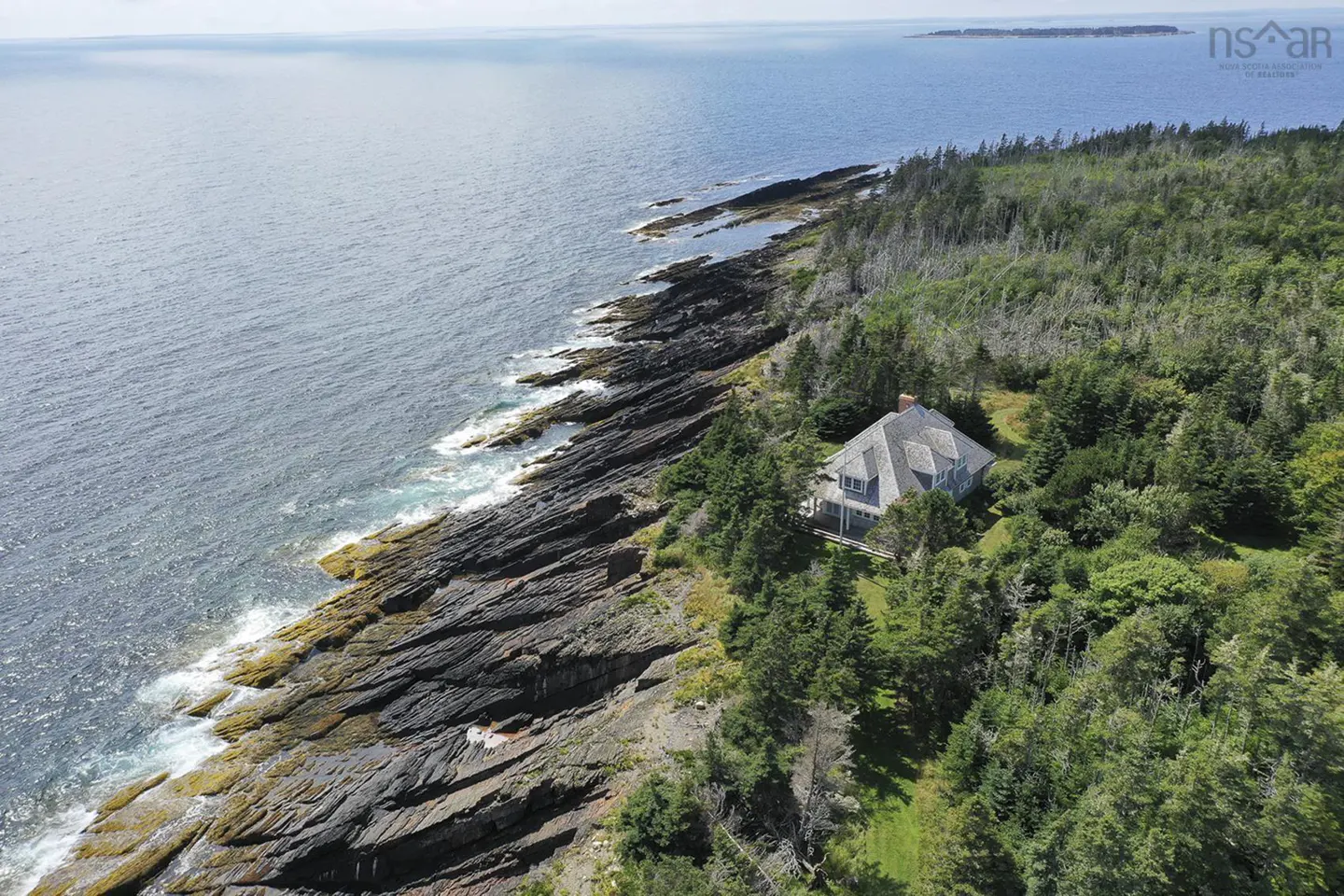Aerial view of a grey house nestled in trees on a rocky coastline next to the blue ocean.