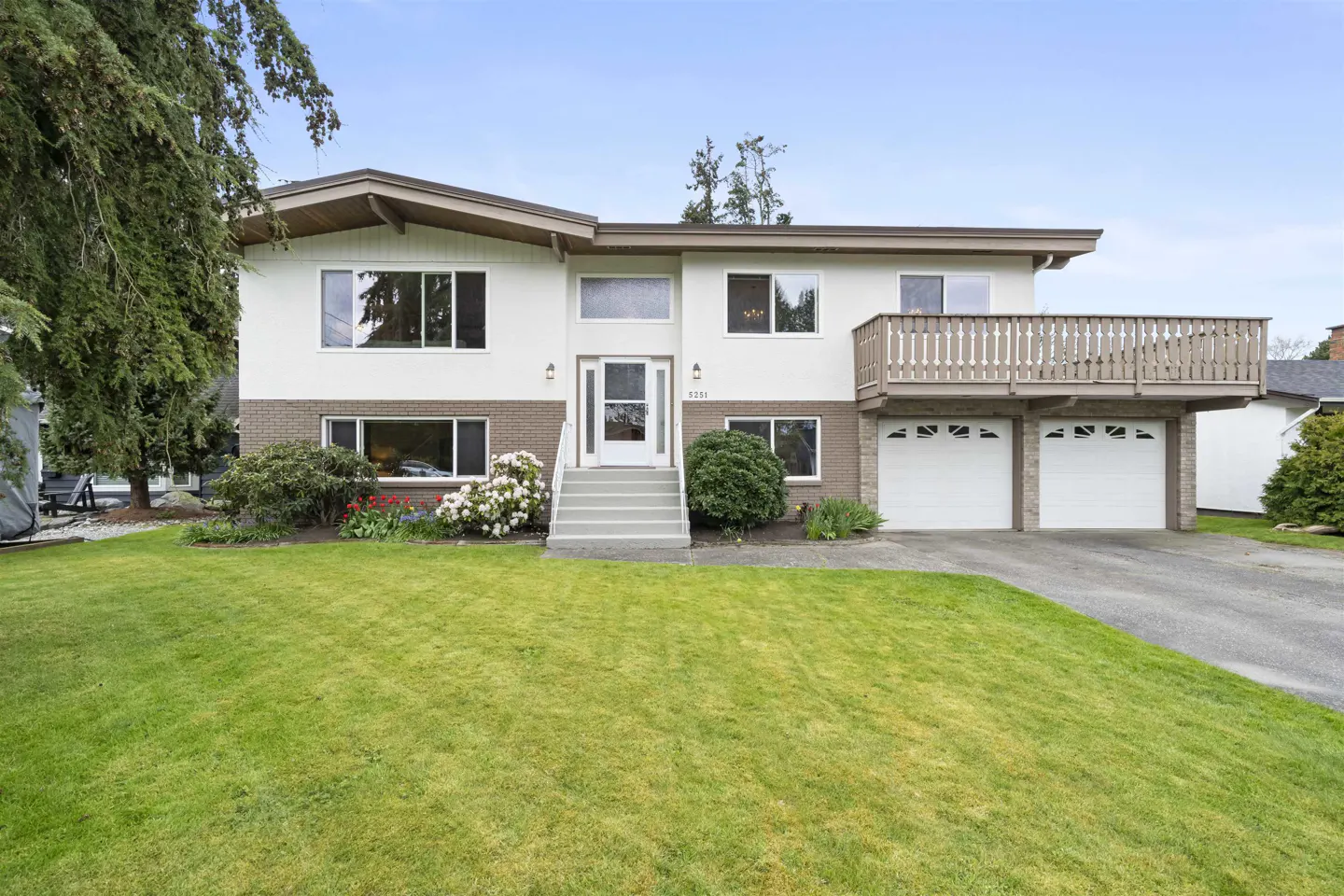 Two-story house with white siding, brick accents, and a brown roof. A green lawn and flower beds are in front. A deck is above the garage.