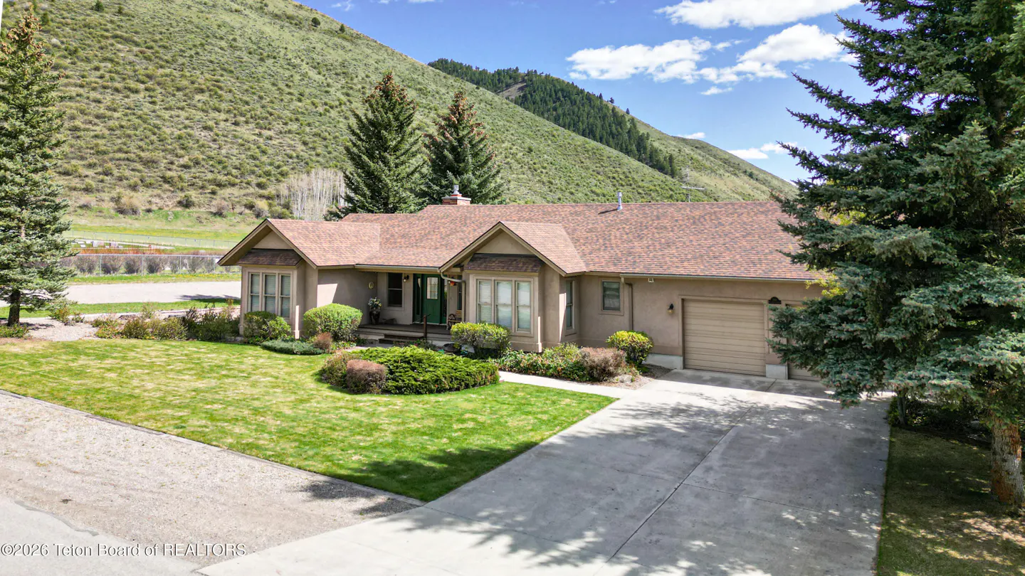 Tan house with brown roof, green door, and garage, set against a grassy mountain backdrop.
