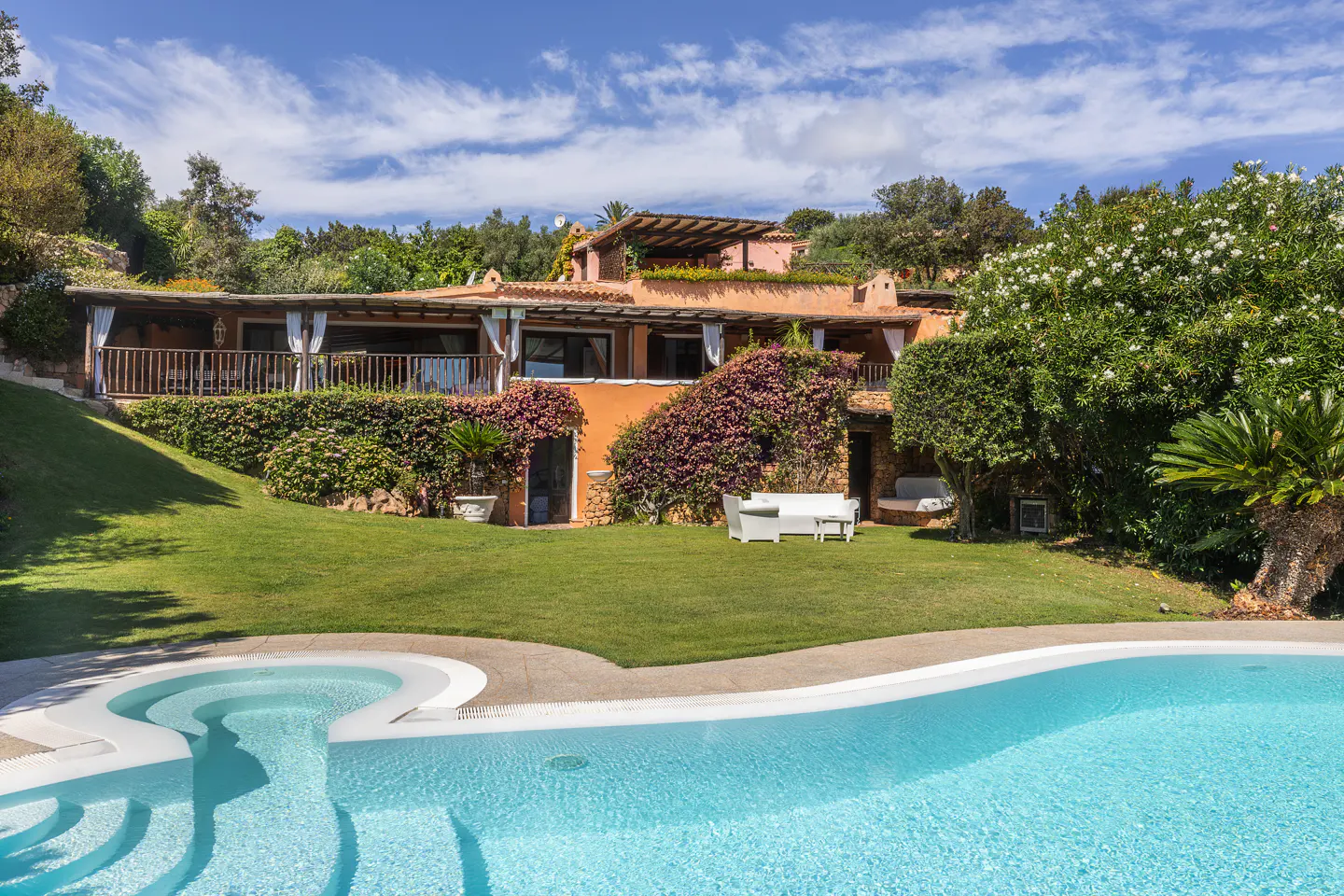 Exterior view of a terracotta-colored house with a pool, green lawn, and flowering plants under a blue sky.