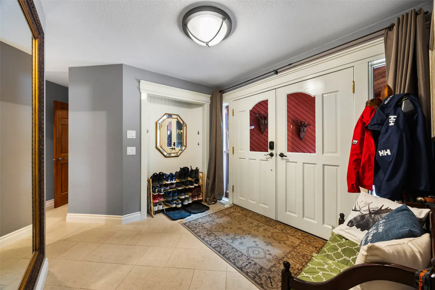 Entryway with beige tile floor, gray walls, and white double doors. A shoe rack sits near an octagonal mirror. A bench with jackets is to the right.