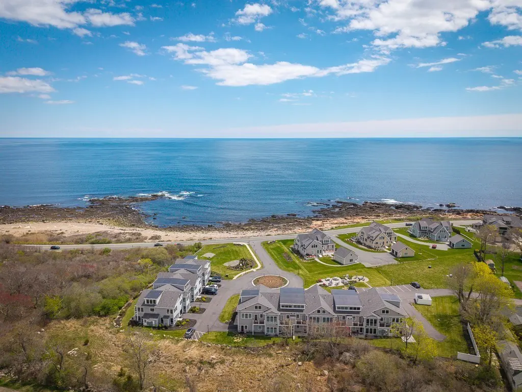 Aerial view of gray condos near the ocean under a blue sky with scattered clouds.