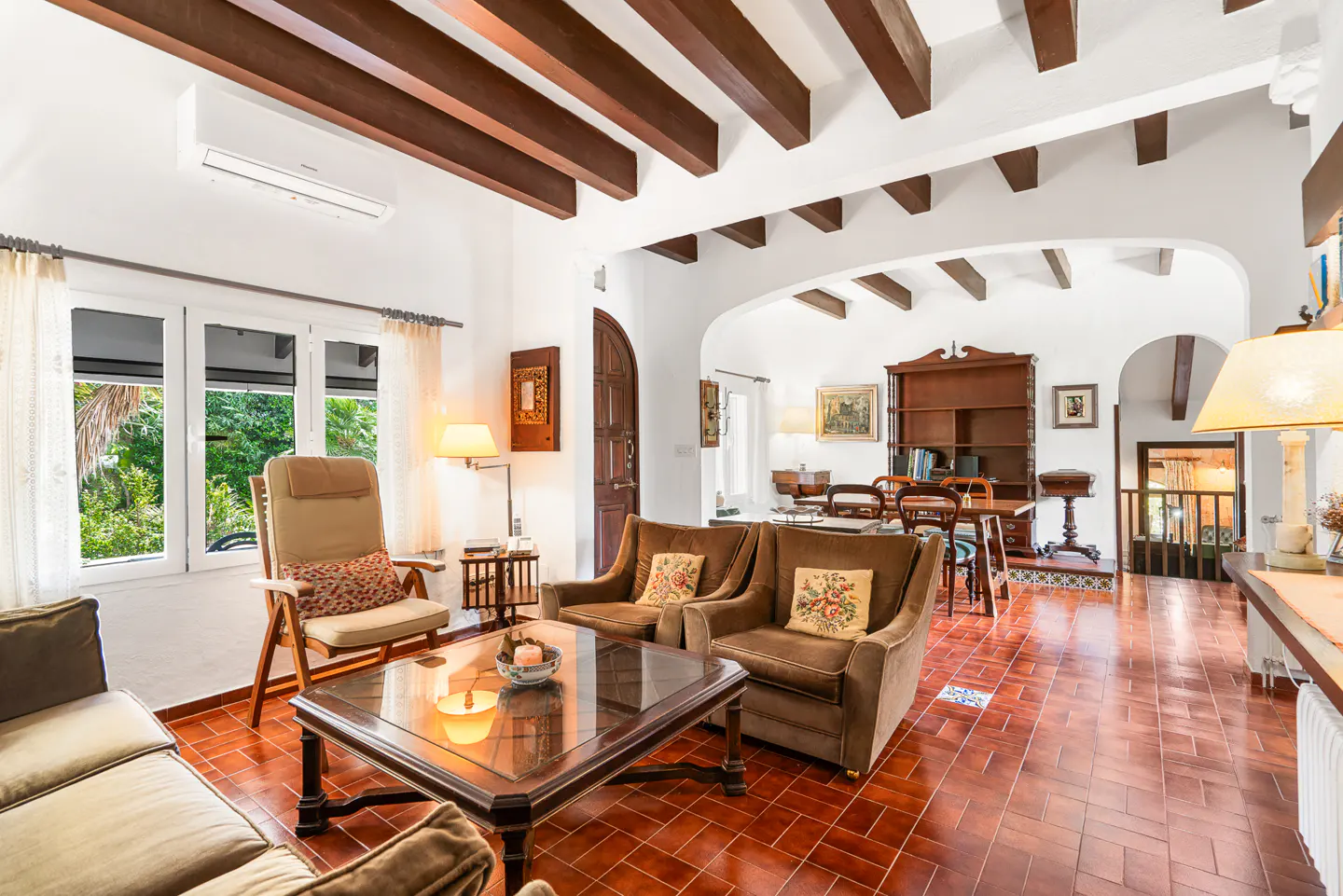 Living room with brown furniture, red tile floor, and white walls. Beamed ceiling and arched doorway lead to dining area.