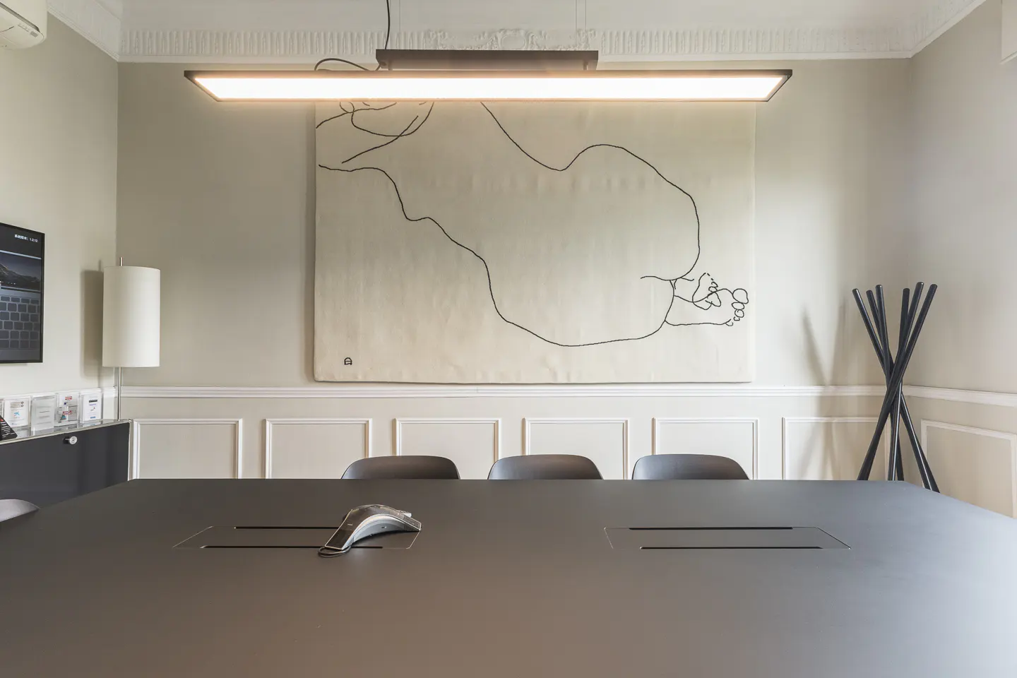 Modern conference room with a large black table, minimalist line-art wall hanging, and a sleek rectangular light fixture.