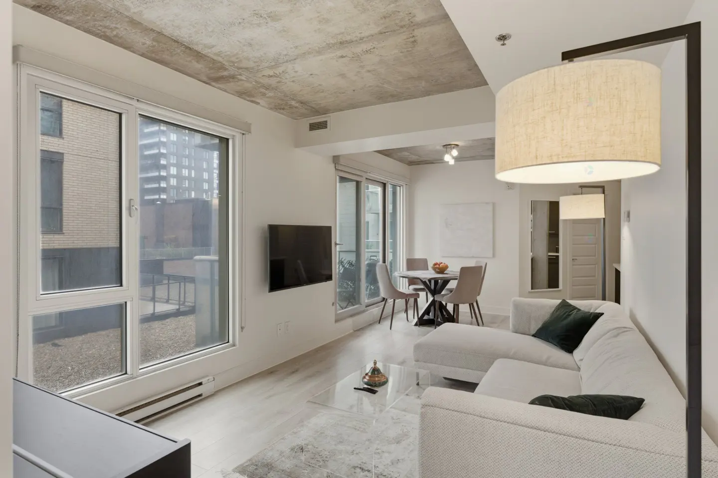 Bright living room with white sectional sofa, floor lamp, and large windows. A dining table and chairs are visible in the background.