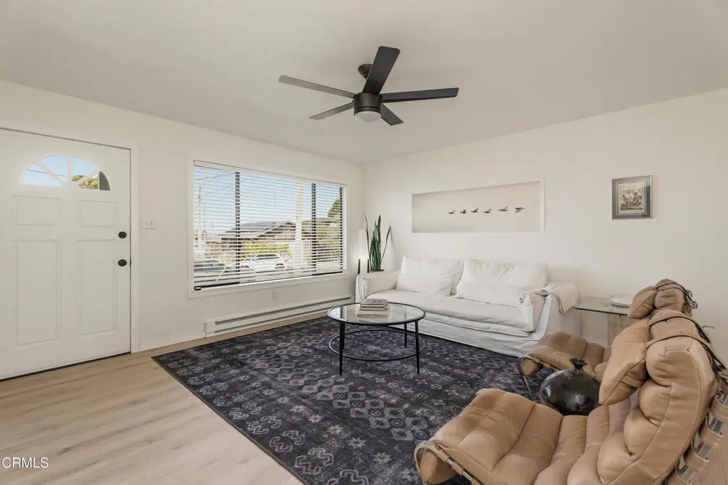 Bright living room with white walls, a black ceiling fan, and a large window. A white sofa sits on a patterned rug, with tan chairs nearby.