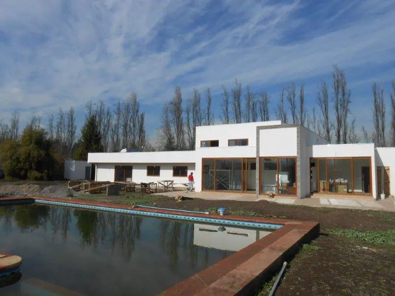 Modern white house with wood trim, a pool with brown brick trim, and a blue sky with trees in the background.