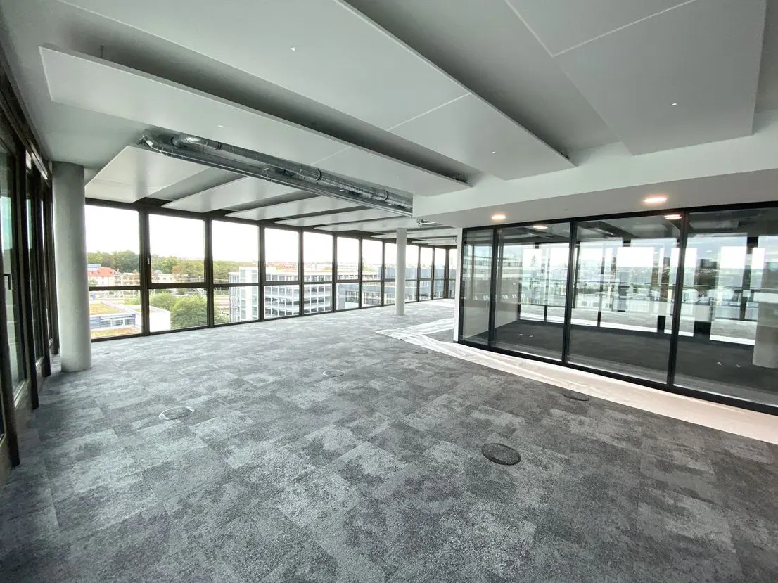 Empty office space with gray carpet, black framed windows, and a view of buildings and trees outside.