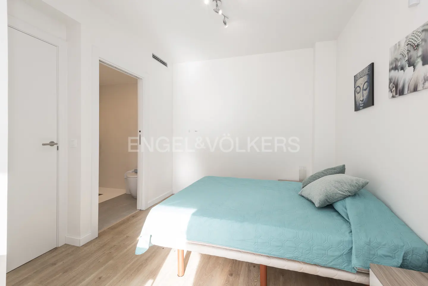 Bright bedroom with a blue bedspread, white walls, and light wood floors. Bathroom visible through an open door. Buddha art on the wall.