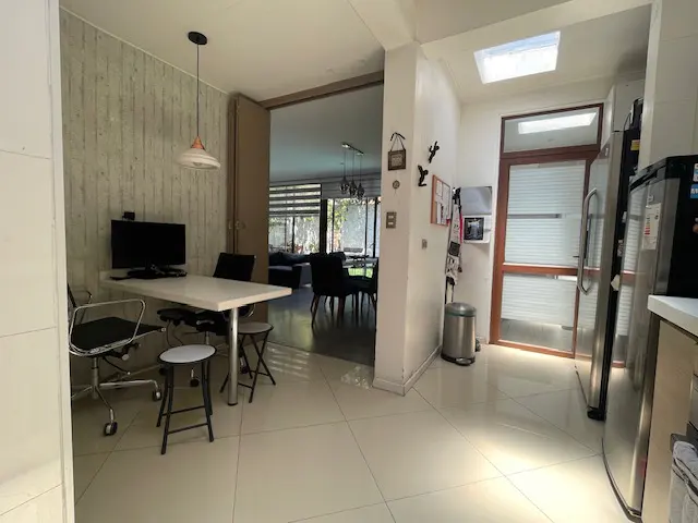 A bright, modern kitchen with white tile floors, a white table with black chairs, and stainless steel appliances. A doorway leads to a dining area.