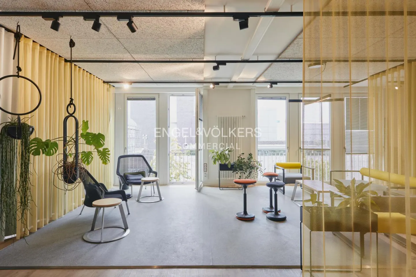 Bright, modern office space with yellow curtains, black track lighting, and hanging plants. Mesh chairs and stools provide seating.