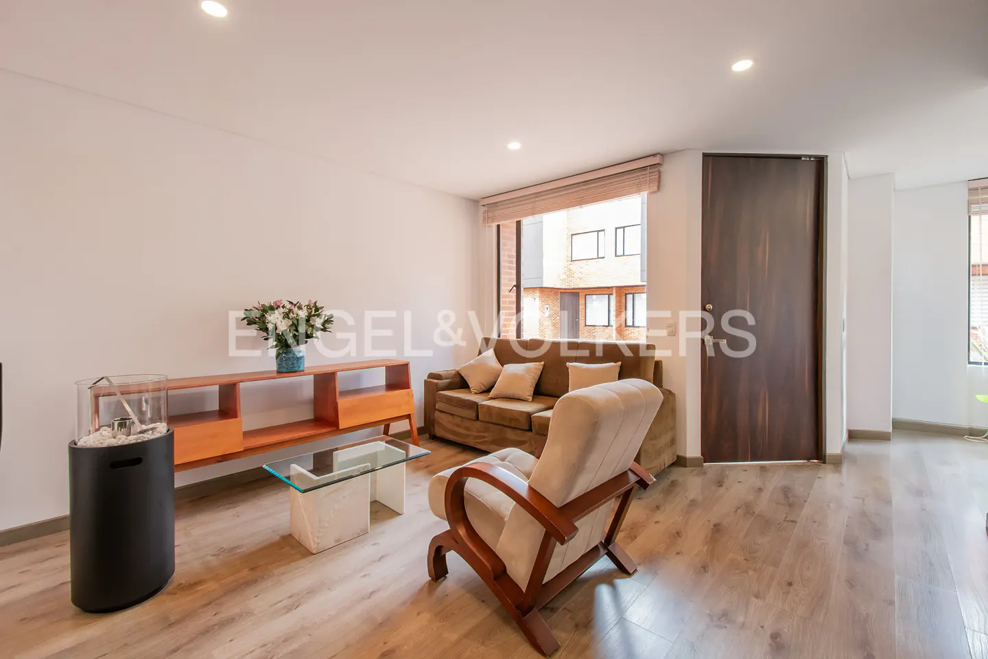 A bright living room with a brown sofa, beige armchair, wood floors, and a glass coffee table. A wooden console table holds flowers.