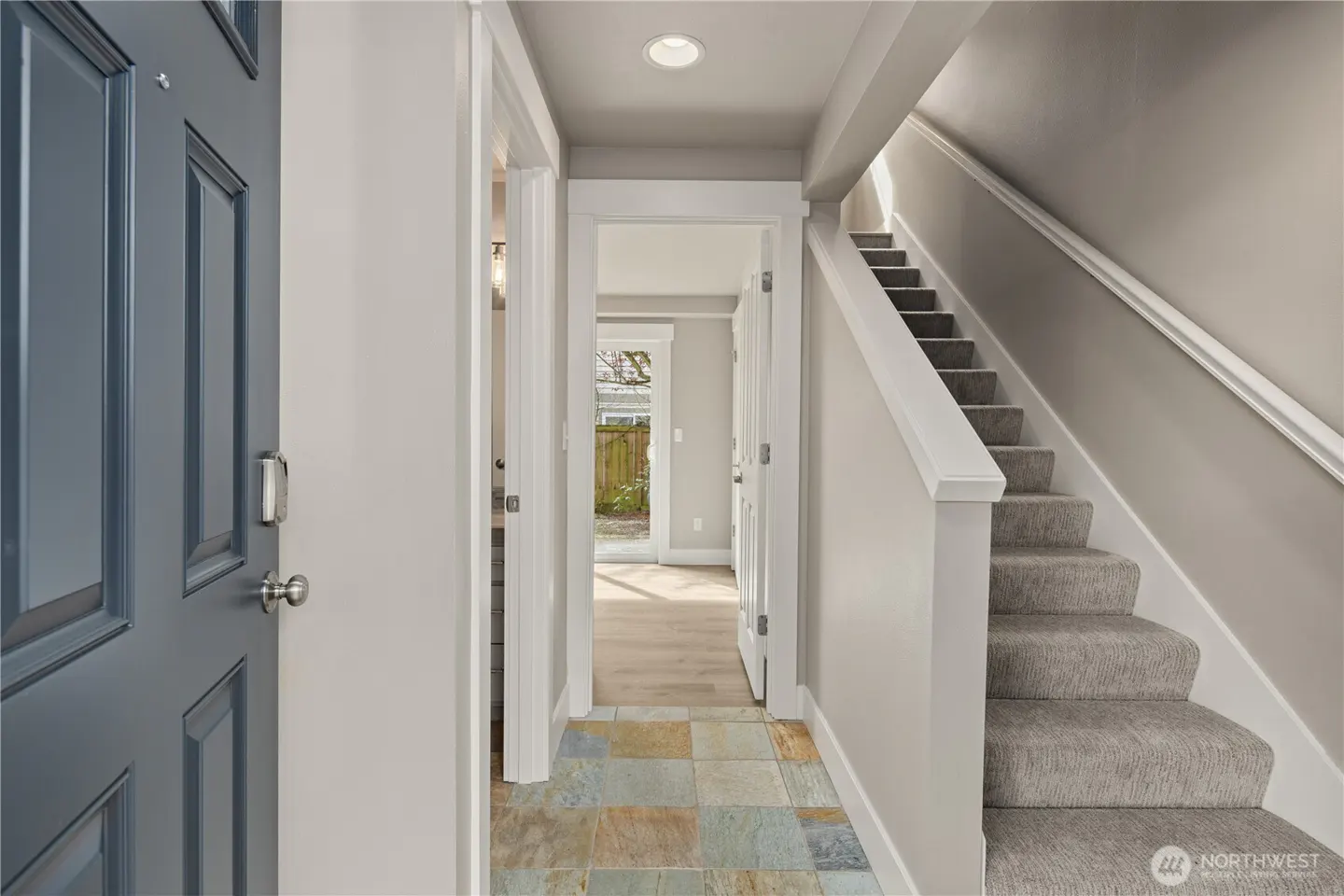Entryway with a blue front door, tiled floor, and carpeted stairs leading up. A doorway leads to a bright room with a view of the backyard.